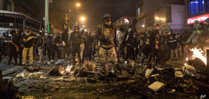 Piles of charred trash burn and smolder in a lamplit city street. Masked community members, illuminated by the fires and LED business signs, walk through the debris as members of the press document the scene.